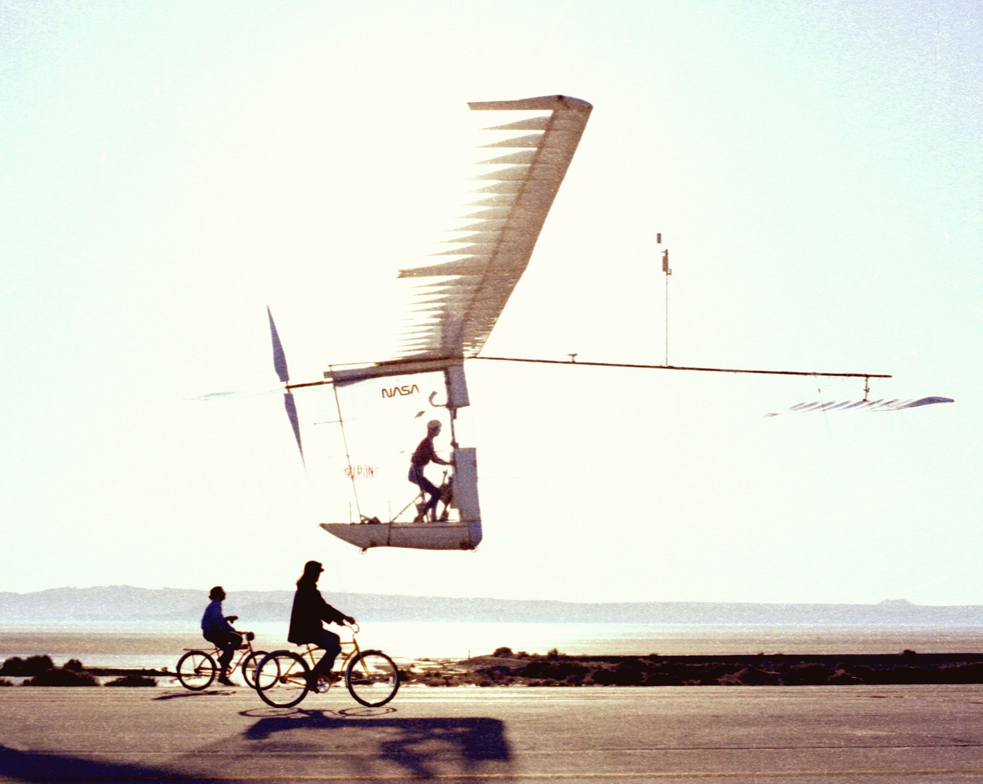 Un avión de pedales casi transparente, de alas larguísimas, vuela a pocos metros del suelo sobre un lago seco mientras dos ciclistas lo siguen por debajo.