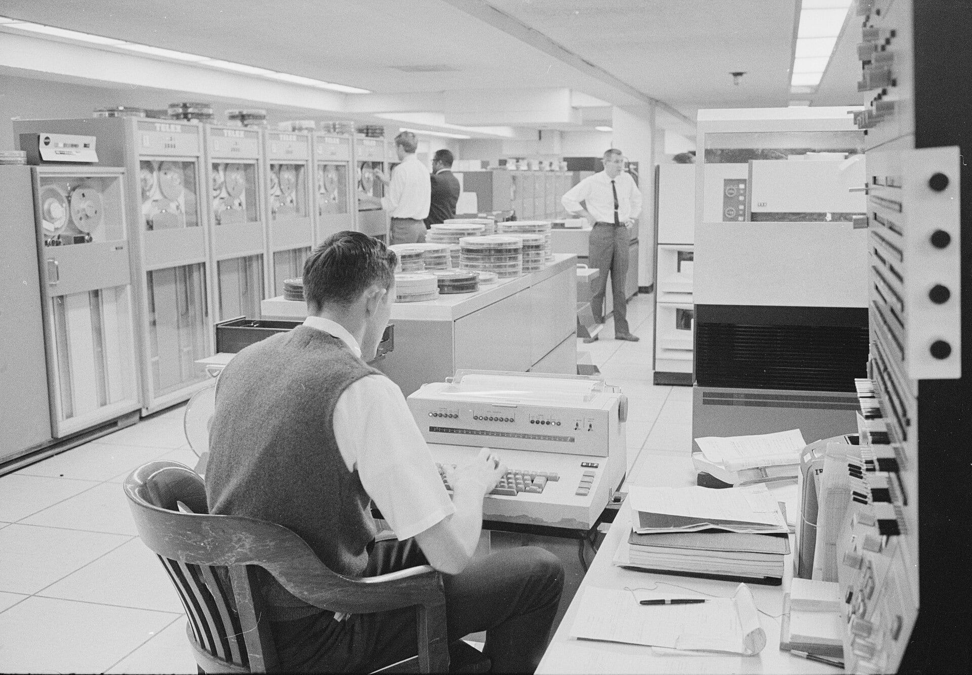 Un operador sentado ante la consola de un ordenador IBM, con armarios de cinta magnética detrás.