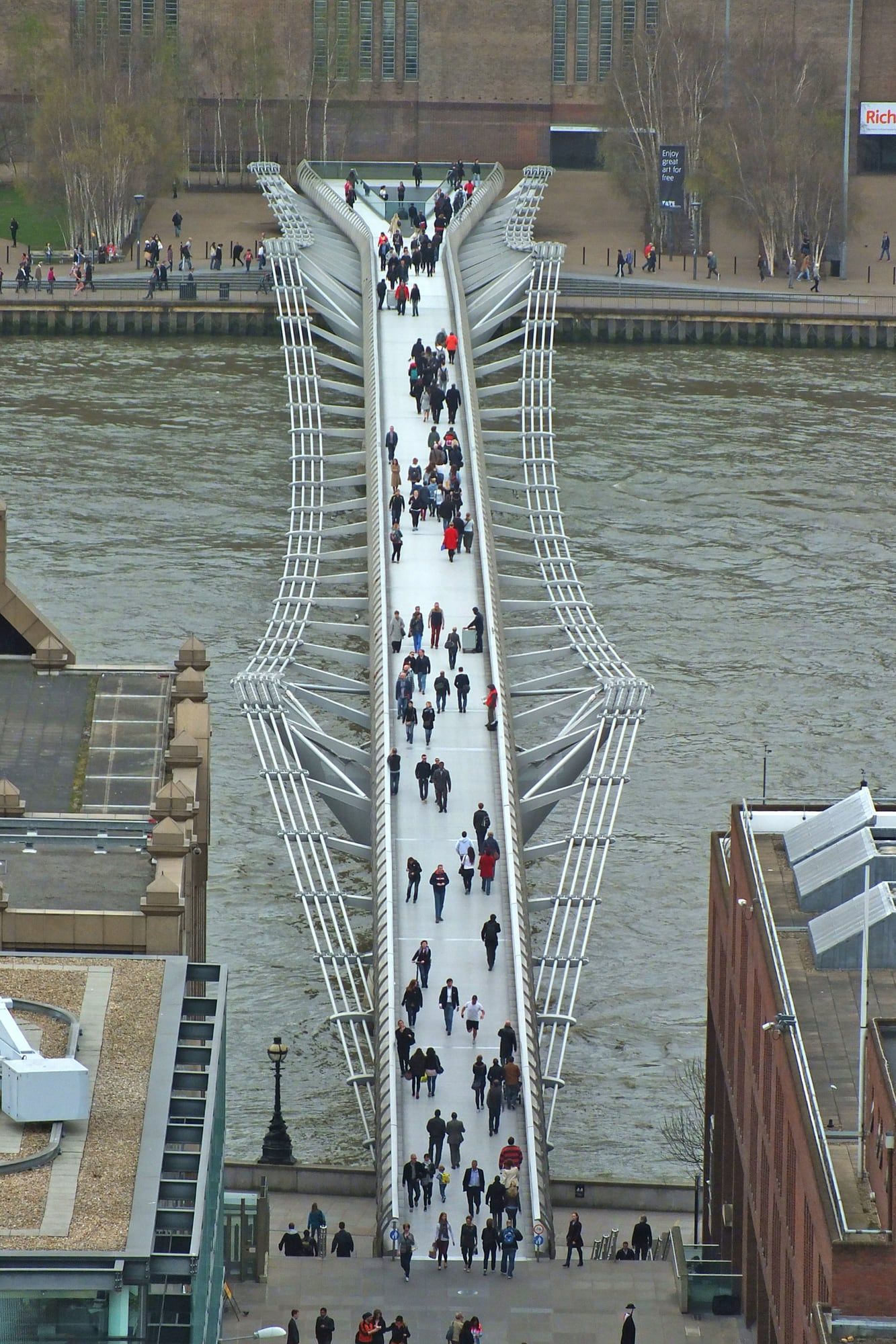Vista cenital de un puente peatonal estrecho sobre el Támesis, lleno de gente.